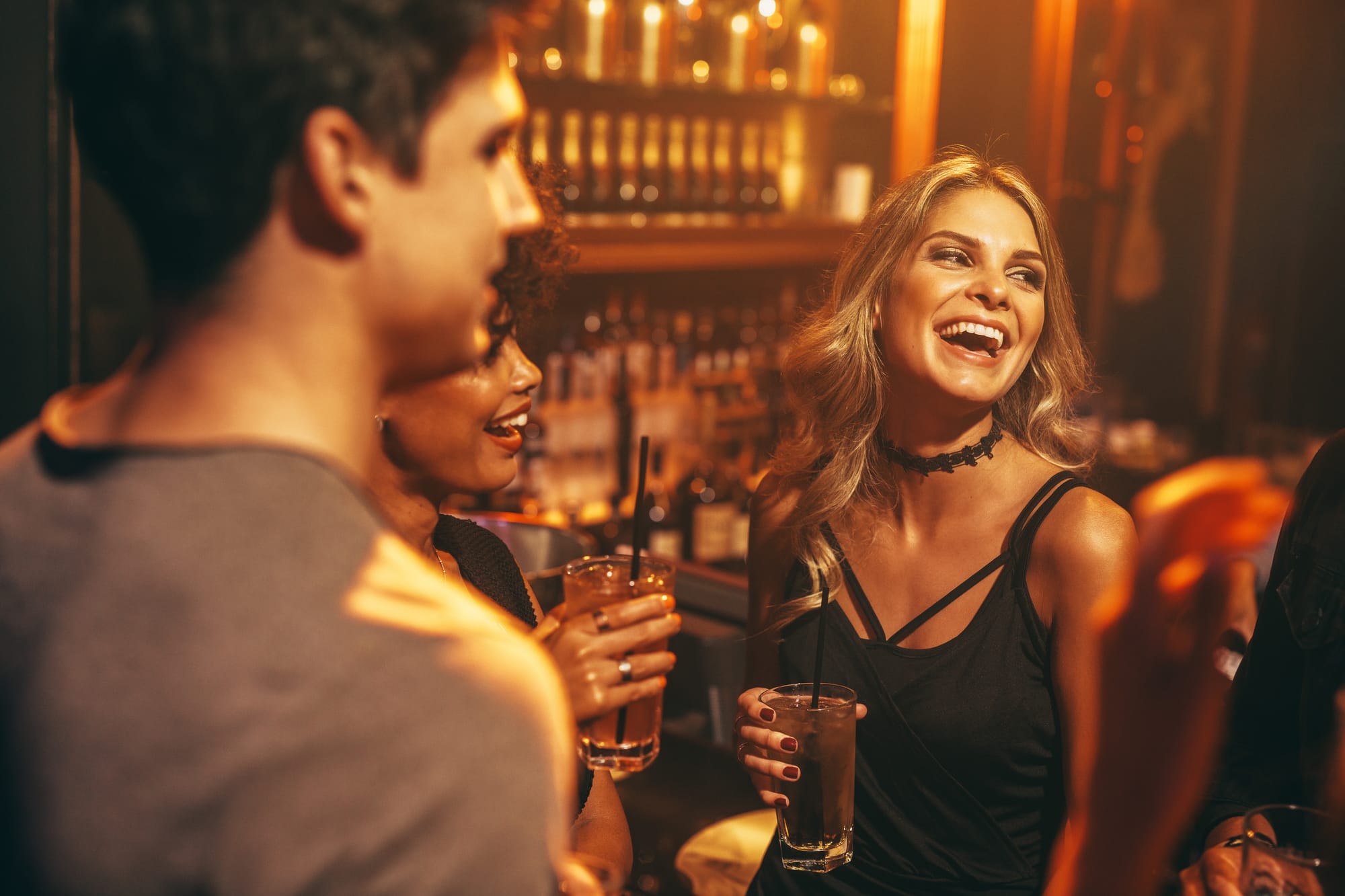 Woman in a bar laughing and smiling with a drink while others watch and listen to the conversation.