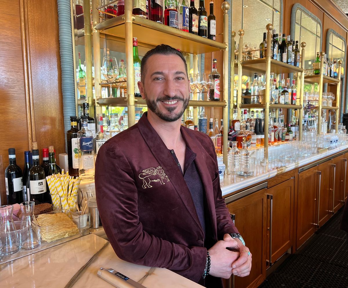 A man is smiling in front of a bar filled with drinks and empty glasses.