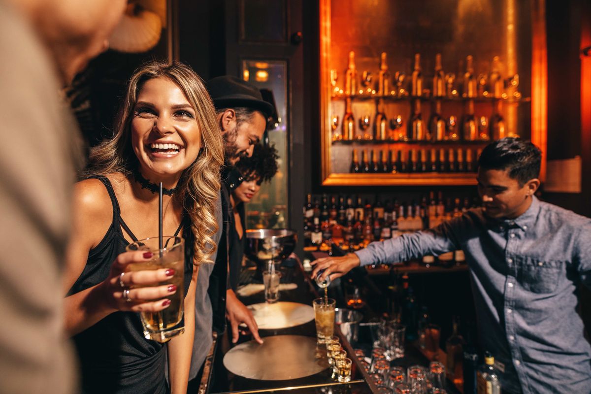 Bartender pours a drink while a woman talks to people at the bar.